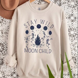 K&ouml;nnte beinhalten: Ein beiger Pullover mit einem grafischen Aufdruck von Skeletten, die um ein Lagerfeuer tanzen, mit dem Text "Stay Wild Moon Child".