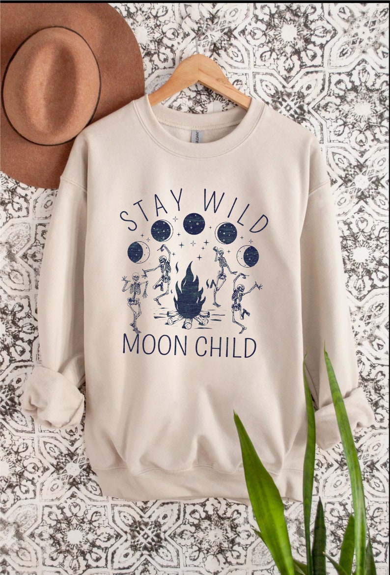 K&ouml;nnte beinhalten: Ein beiger Pullover mit einem grafischen Aufdruck von Skeletten, die um ein Lagerfeuer tanzen, mit dem Text "Stay Wild Moon Child".