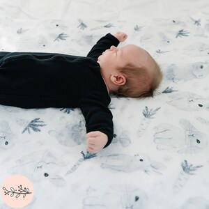 May include: A baby wearing a black long-sleeved onesie is lying on a white muslin swaddle blanket with a watercolor print of bunnies and carrots.