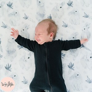 May include: A baby wearing a black long-sleeved onesie lies on a white blanket with a watercolor bunny print. The baby has their eyes closed and is sleeping peacefully.