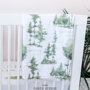 May include: A white muslin baby blanket with a watercolor forest print. The blanket features green trees and the text "GREYCEN isaiah" repeated throughout.