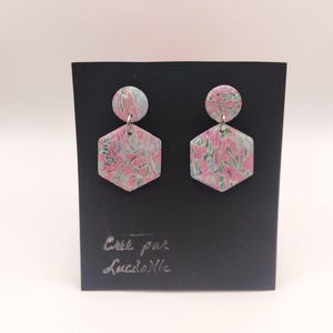 Op de afbeelding: Een paar hexagonale oorbellen met een roze en groen bloemenpatroon en een klein rond bovenstuk. De oorbellen worden gepresenteerd op een zwarte kaart met de tekst "Cree par Lucile" in zilver.