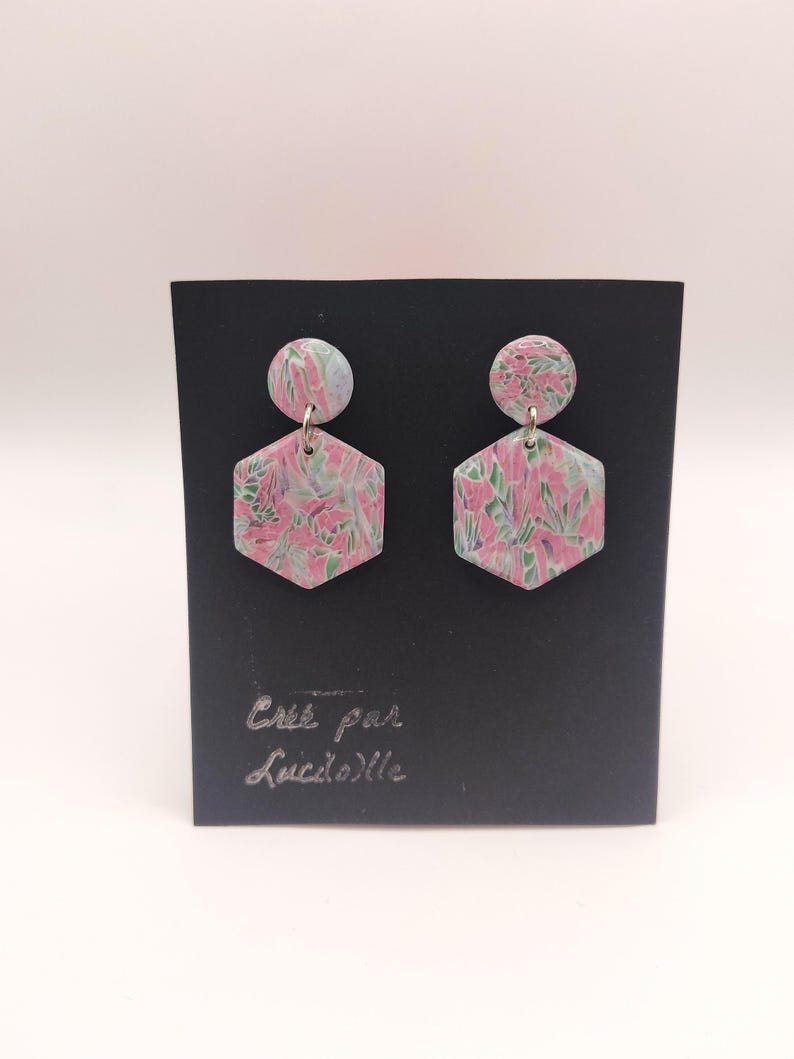 Op de afbeelding: Een paar hexagonale oorbellen met een roze en groen bloemenpatroon en een klein rond bovenstuk. De oorbellen worden gepresenteerd op een zwarte kaart met de tekst "Cree par Lucile" in zilver.