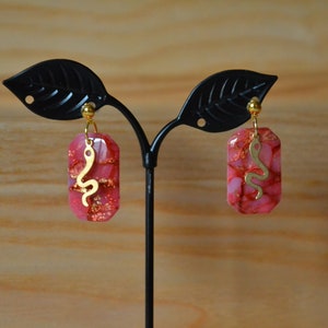 Peut inclure: Une paire de boucles d'oreilles roses et dor&eacute;es avec un motif de serpent. Les boucles d'oreilles sont en r&eacute;sine et ont un charme de serpent dor&eacute;.