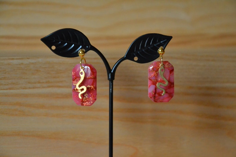 Peut inclure: Une paire de boucles d'oreilles roses et dor&eacute;es avec un motif de serpent. Les boucles d'oreilles sont en r&eacute;sine et ont un charme de serpent dor&eacute;.