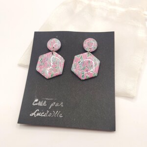 Op de afbeelding: Een paar roze en groene zeshoekige oorbellen met een bloemenpatroon. De oorbellen worden gepresenteerd op een zwarte kaart met de tekst "Cree par Luciole".