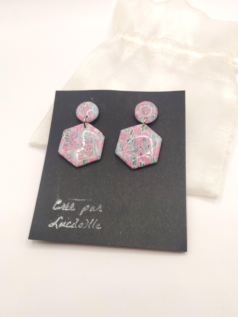 Op de afbeelding: Een paar roze en groene zeshoekige oorbellen met een bloemenpatroon. De oorbellen worden gepresenteerd op een zwarte kaart met de tekst "Cree par Luciole".