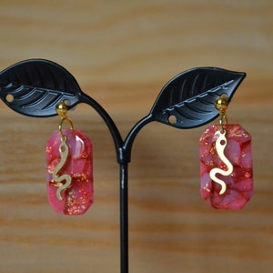 Peut inclure: Une paire de boucles d'oreilles roses et dor&eacute;es avec un motif de serpent. Les boucles d'oreilles sont en r&eacute;sine et ont une finition dor&eacute;e.