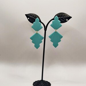 Peut inclure: Une paire de boucles d'oreilles g&eacute;om&eacute;triques bleu turquoise avec des accents dor&eacute;s sur un support de boucles d'oreilles noir.