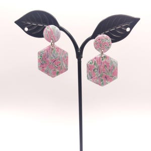 Op de afbeelding: Een paar roze en groene oorringen met een bloemenpatroon en zilveren haakjes. De oorringen worden getoond op een zwarte oorringstandaard.