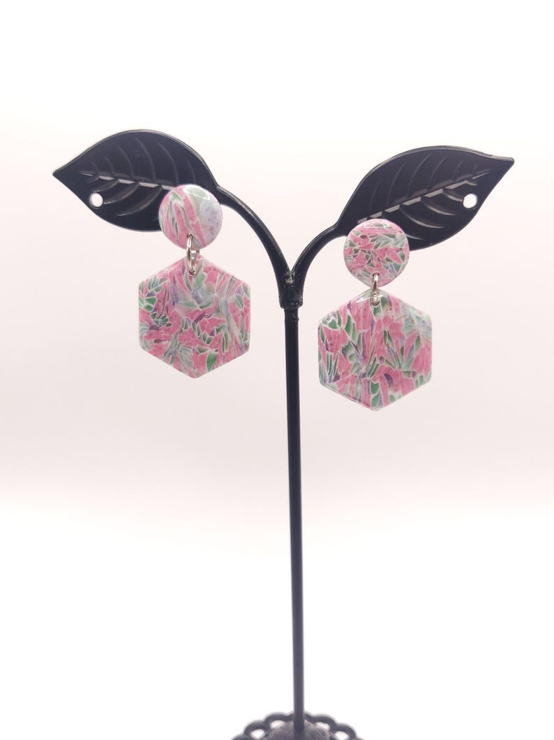 Op de afbeelding: Een paar roze en groene oorringen met een bloemenpatroon en zilveren haakjes. De oorringen worden getoond op een zwarte oorringstandaard.