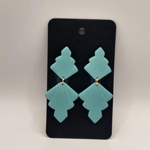 Peut inclure: Une paire de boucles d'oreilles g&eacute;om&eacute;triques bleu turquoise avec des accents dor&eacute;s. Les boucles d'oreilles sont pr&eacute;sent&eacute;es sur une carte noire.