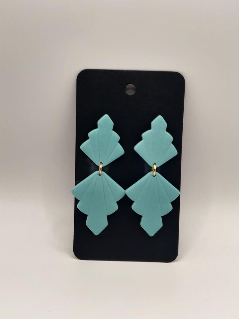 Peut inclure: Une paire de boucles d'oreilles g&eacute;om&eacute;triques bleu turquoise avec des accents dor&eacute;s. Les boucles d'oreilles sont pr&eacute;sent&eacute;es sur une carte noire.