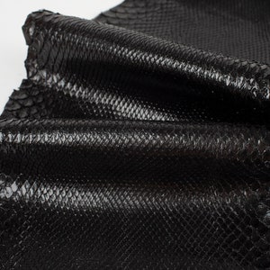 Puede incluir: Cuero de serpiente negro con una superficie texturizada y escamosa. El cuero está doblado y muestra la veta natural de la piel.