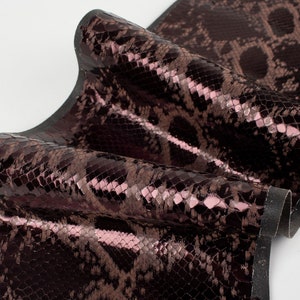 May include: Close-up of a dark brown snakeskin fabric with a subtle pink sheen. The fabric is folded and has a black backing.