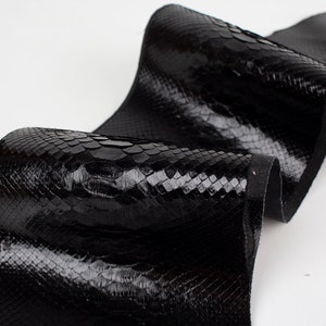 以下が含まれることがあります： 光沢のある黒いヘビ革。鱗模様があり、カールして折りたたまれており、裏側が見えています。アクセサリーや革製品の製作に適した素材です。