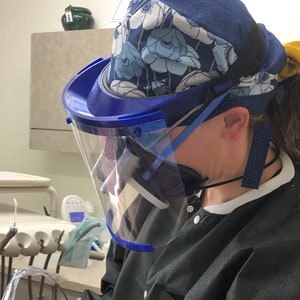 May include: A dentist wearing a blue surgical cap with a floral pattern, a clear face shield, and a black scrub top, is working on a patient's teeth.