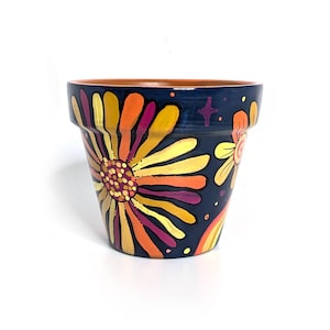 May include: A hand-painted terracotta flower pot with a dark blue background. The pot features a large, colorful daisy design in shades of yellow, orange, and purple. Additional floral and rainbow accents are also present.