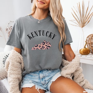 Peut inclure: Un t-shirt gris avec un motif noir représentant l'état du Kentucky avec un imprimé léopard rose et noir. Le texte "Kentucky" est au-dessus du contour de l'état.