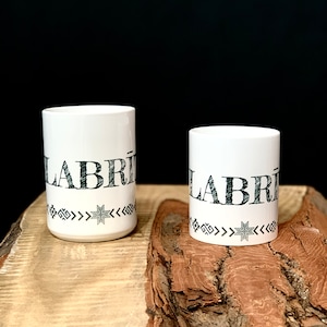 May include: Two white ceramic mugs with black lettering that reads "LABRI" and a black and white geometric design around the bottom. The mugs are sitting on a wooden surface.