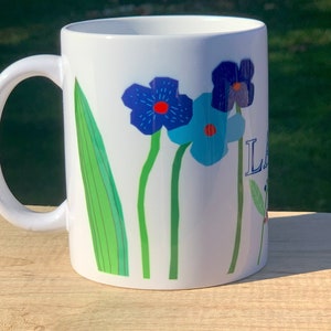 May include: White ceramic mug with a floral design featuring blue and green flowers. The mug has a white handle and the text "LA" is printed on the side.