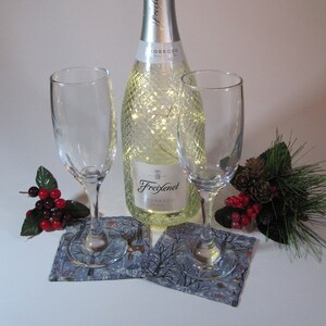 May include: Two champagne flutes and a bottle of Freixenet Prosecco with a string of lights inside. The bottle and glasses are on blue and white patterned coasters with a winter scene.
