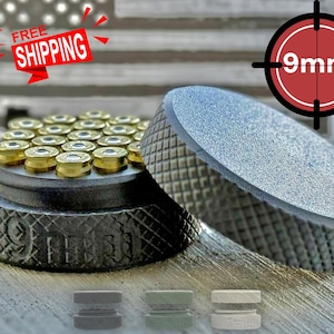 May include: A dark gray 9mm ammunition container with a textured surface, holding several brass-colored bullets. The lid is open, revealing the contents. The container has the text "9mm" engraved on the side. A red target with "9mm" is in the background.
