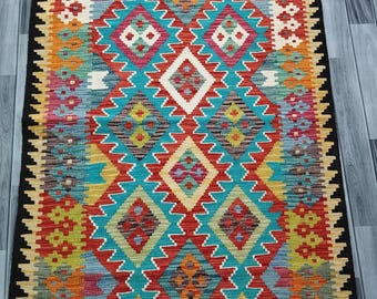 May include: A rectangular, hand-woven rug with a vibrant geometric pattern. The rug features a black border and a central design of diamonds and zigzags in turquoise, red, yellow, and orange. The rug is likely made of wool.