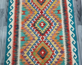 May include: A colorful rug with a geometric pattern in shades of red, orange, yellow, green, blue, and purple. The rug has a teal border with white accents and fringe on the ends.