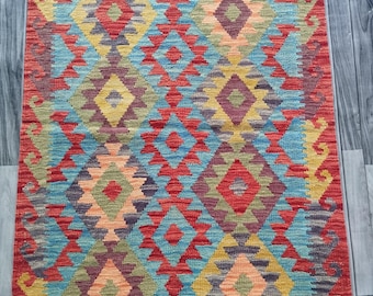 May include: A rectangular, hand-woven rug with a vibrant geometric pattern. The rug features a red border and a central design of diamonds and triangles in shades of red, blue, yellow, and orange. The rug has a fringed edge.