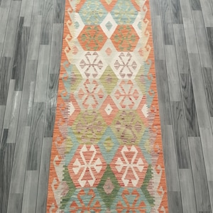 May include: A rectangular, patterned rug with a geometric design. The rug features a combination of colors, including orange, green, blue, and cream. The design includes diamond and snowflake-like shapes. The rug is placed on a gray wooden floor.