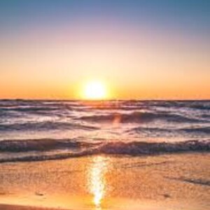 Könnte beinhalten: Ein Foto eines Strandes bei Sonnenuntergang. Der Himmel wechselt von tiefblau zu orange und gelb in der Nähe des Horizonts. Die Sonne ist eine helle Kugel, die sich in den Meereswellen und im nassen Sand spiegelt.