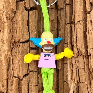 Może przedstawiać: Zielono-żółty brelok z figurką Lego Krusty'ego Klauna. Figurka ma różową koszulę, niebieskie włosy i brązowy nos.