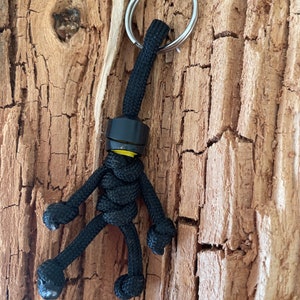 Puede incluir: Un llavero de paracord negro con una cabeza amarilla y un anillo de llave plateado. El llavero tiene forma de figura de palo.