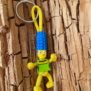 May include: A yellow paracord keychain with a blue hair and yellow body figure attached. The figure has a green shirt and red beads around its neck.