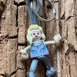 Puede incluir: Un llavero de paracord gris y blanco con una peque&ntilde;a figura de pl&aacute;stico gris de una mujer con cabello rubio y una blusa con estampado azul y rosa. La figura est&aacute; unida al llavero con un anillo plateado.