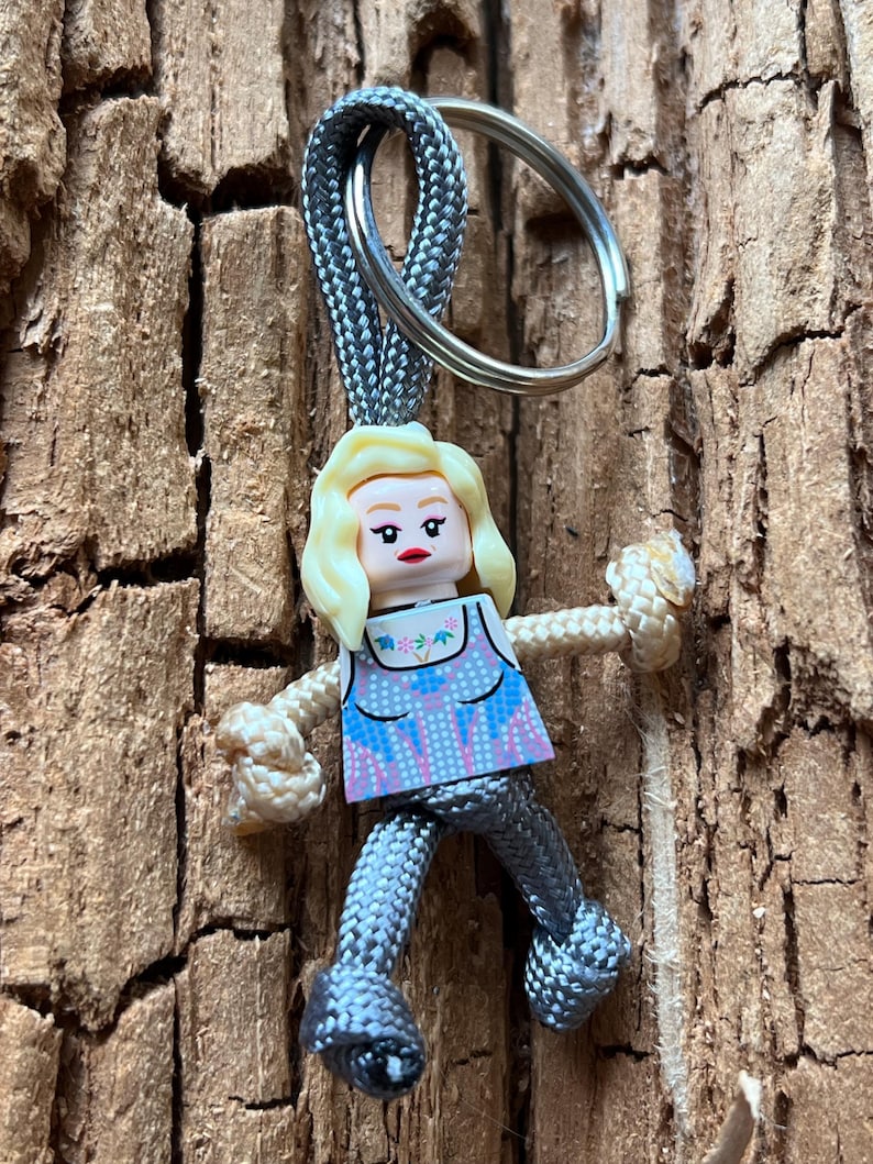 Puede incluir: Un llavero de paracord gris y blanco con una peque&ntilde;a figura de pl&aacute;stico gris de una mujer con cabello rubio y una blusa con estampado azul y rosa. La figura est&aacute; unida al llavero con un anillo plateado.