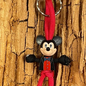 May include: A red and black string figure of Mickey Mouse wearing a tuxedo. The figure is attached to a silver key ring.