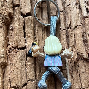 Puede incluir: Un llavero de paracord gris con una peque&ntilde;a figura de una persona escalando una pared. La figura lleva una camisa azul y rosa y tiene el pelo rubio.