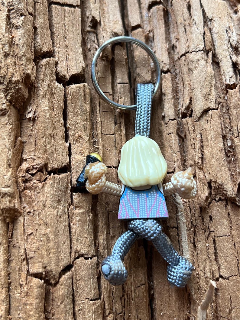 Puede incluir: Un llavero de paracord gris con una peque&ntilde;a figura de una persona escalando una pared. La figura lleva una camisa azul y rosa y tiene el pelo rubio.