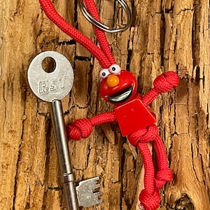 Może przedstawiać: Brelok z czerwonego i pomarańczowego filcu z czerwoną figurką z filcu przypominającą popularną postać z kreskówek. Figurka trzyma srebrny klucz z literami "RST" na górze.