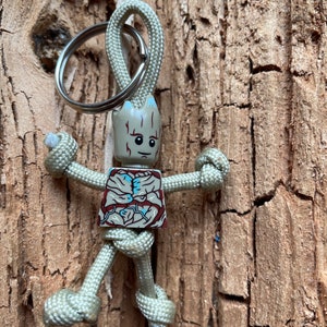 May include: A beige and brown paracord keychain with a small Groot figurine attached. The keychain is tied in a knot to resemble a person's arms and legs.