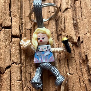 Puede incluir: Un llavero de paracord gris con una peque&ntilde;a figura de Lego de pelo rubio que sostiene un micr&oacute;fono. La figura lleva una blusa con estampado azul y blanco y una falda gris.