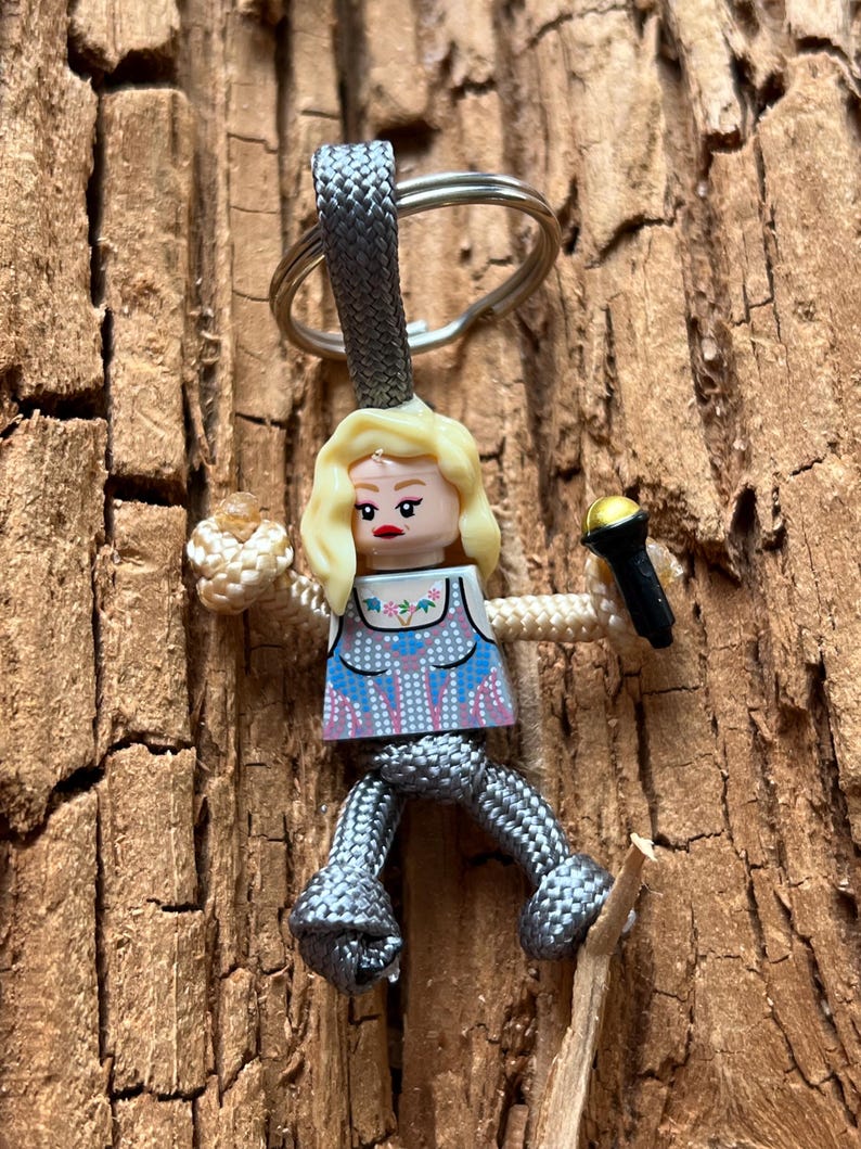 Puede incluir: Un llavero de paracord gris con una peque&ntilde;a figura de Lego de pelo rubio que sostiene un micr&oacute;fono. La figura lleva una blusa con estampado azul y blanco y una falda gris.