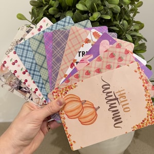 Könnte beinhalten: Ein Fächer aus rechteckigen Karten mit verschiedenen Designs, darunter eine Hello Autumn-Karte mit Kürbissen, ein Erdbeermuster, Karomuster und Kirschillustrationen. Die Karten werden in einem Stapel gehalten und zeigen eine Vielzahl bunter Grafiken.