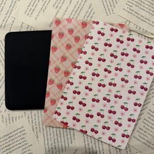 Könnte beinhalten: Ein schwarzer E-Reader, der teilweise von zwei rechteckigen Karten bedeckt ist. Eine Karte zeigt ein rosa-weißes Karomuster mit roten Erdbeeren, während die andere einen weißen Hintergrund mit einem sich wiederholenden Muster aus roten Kirschen und rosa Blüten aufweist.