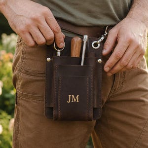 Pode incluir: Uma bolsa de ferramentas em couro castanho escuro com várias ranhuras e bolsos verticais, presa a um cinto com um clipe de metal. O bolso frontal tem as iniciais JM gravadas em dourado. A bolsa contém um cabo de madeira e ferramentas.