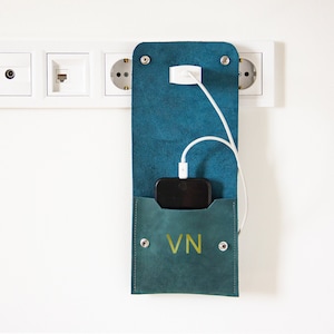May include: A teal leather phone charging pouch with a white charging cable and a black smartphone inside. The pouch has the initials "VN" embossed on the front.