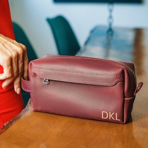 Puede incluir: Una bolsa de tocador de viaje de cuero burdeos con cierre de cremallera y un monograma dorado que dice "DKL".