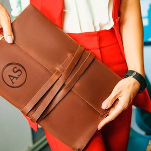 May include: A brown leather portfolio with a flap closure and a strap. The portfolio has a monogrammed "AS" in a circle on the front.